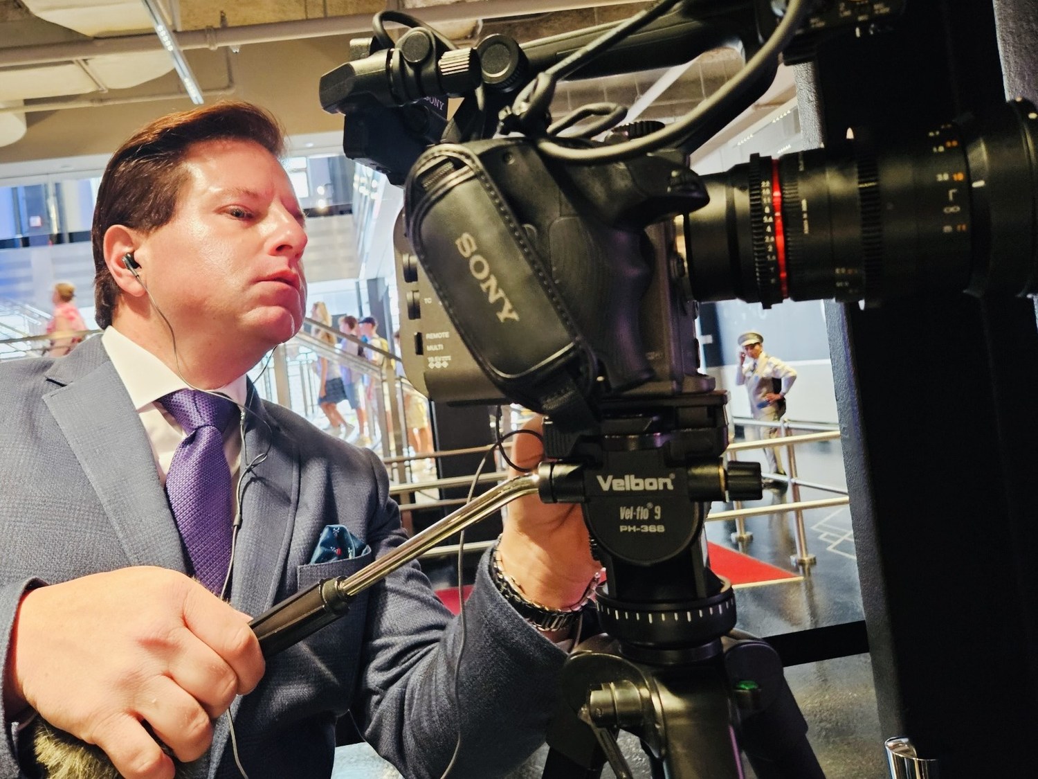 Brian Calfano in a grey suit and earpiece, crouched behind a tripod-mounted Sony video camera while setting up a shot in a busy public concourse.