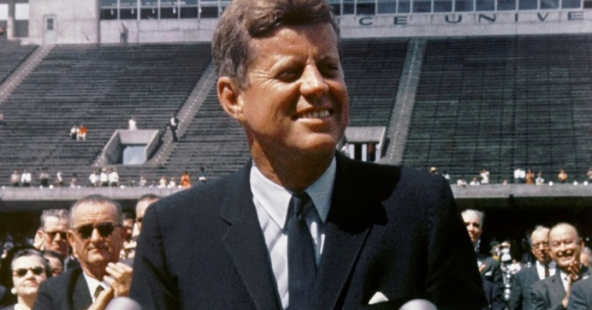 President John F. Kennedy smiling at microphones, Rice University's stadium seating and a campus building reading '...CE UNIVE...' visible behind him, during his 'We choose to go to the Moon' address, September 12, 1962.